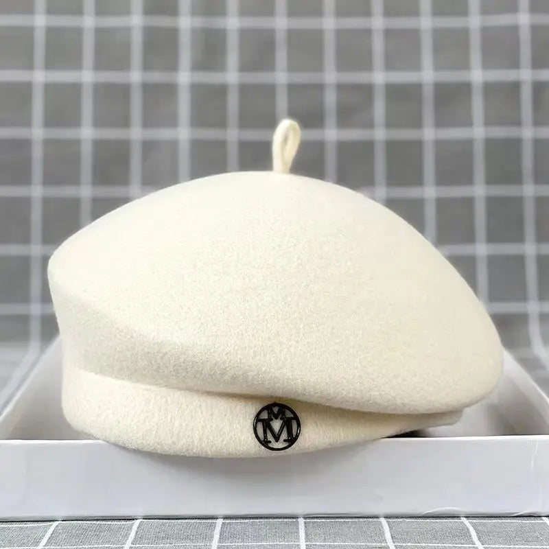 Cream wool felt beret with a small stem top and a circular black metallic logo detail on the brim.
