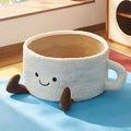 White plush coffee-cup-shaped cat bed with handle, smiling face embroidery, brown corduroy feet, and textured inner bowl.