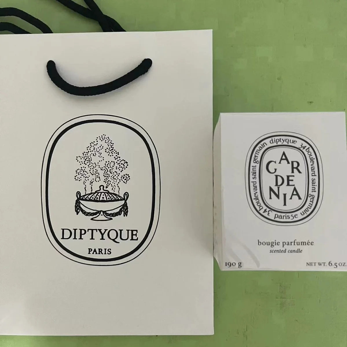 Gardenia scented candle in a white box with black branding, next to a white paper gift bag on a green background.