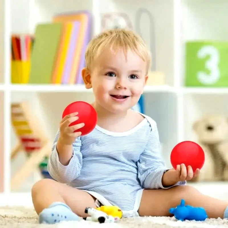 A smiling toddler sitting on a rug and holding two bright red plastic balls.