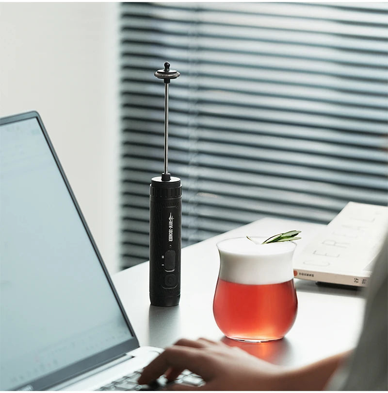Black handheld electric milk frother with a metal whisk wand, placed on a desk next to a glass of foam-topped tea.