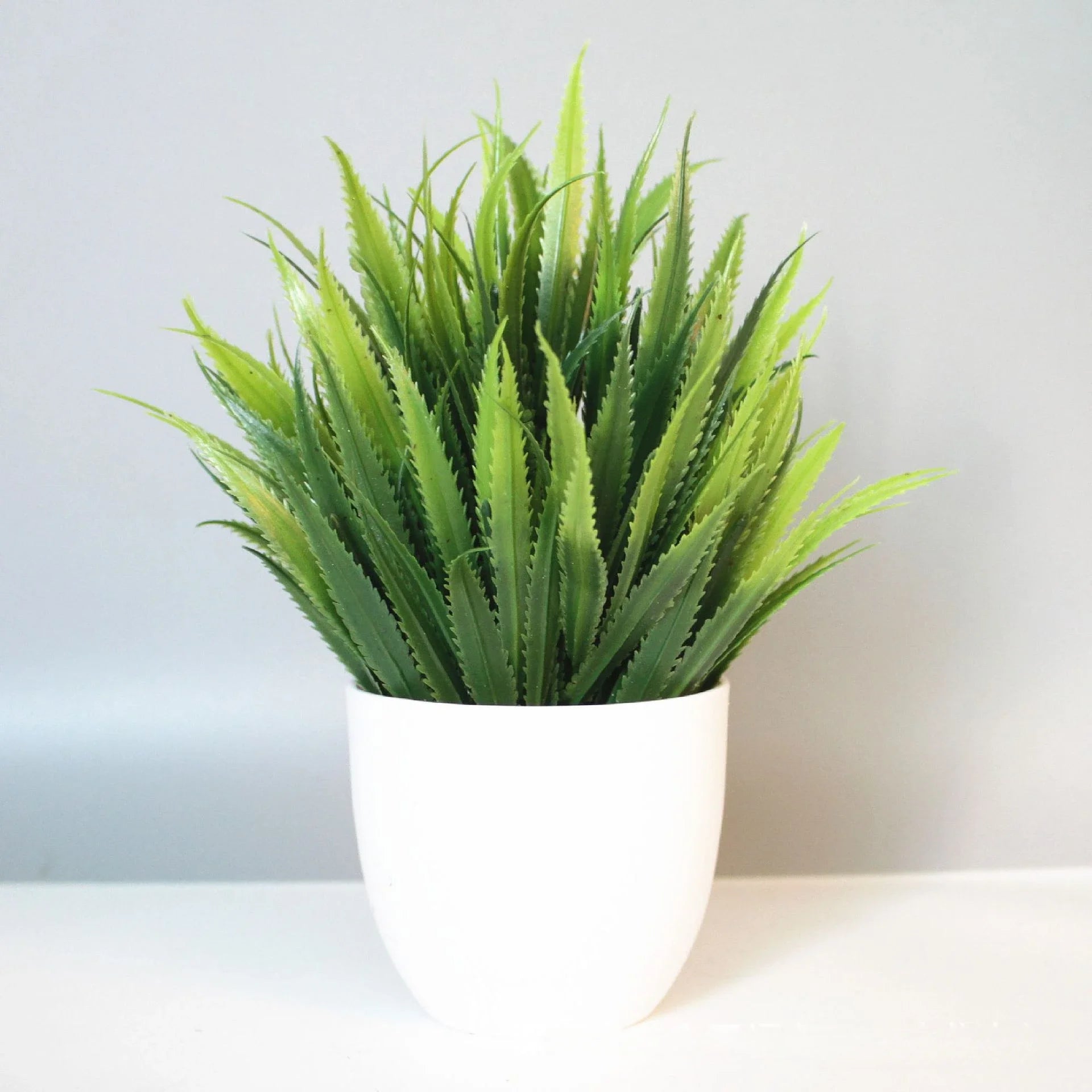 Artificial spiky green grass plant in a simple, solid white ceramic-style pot against a plain gray background.