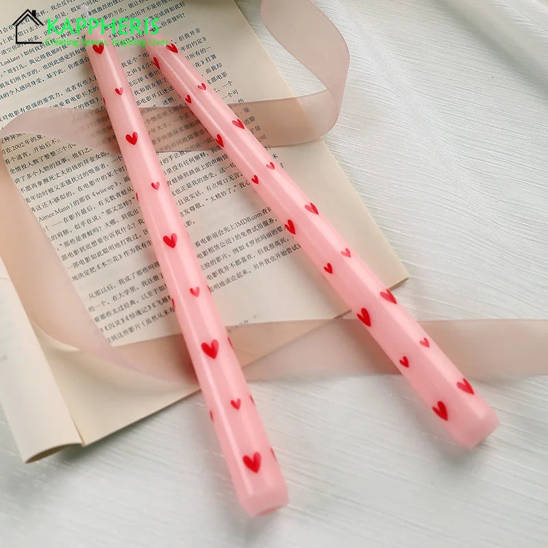 Pair of light pink taper candles decorated with a repeating pattern of small red hearts, resting on book pages.