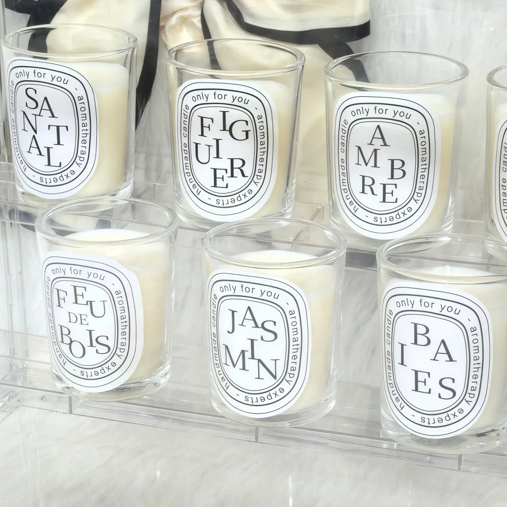 Set of six scented aromatherapy candles in clear glass jars with minimalist white labels on a clear tiered stand.
