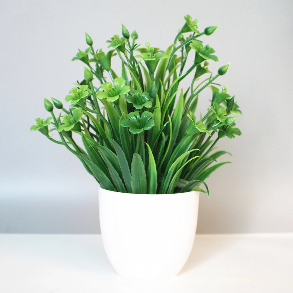 Artificial green plant with small flowers in a simple white pot against a neutral gray background.