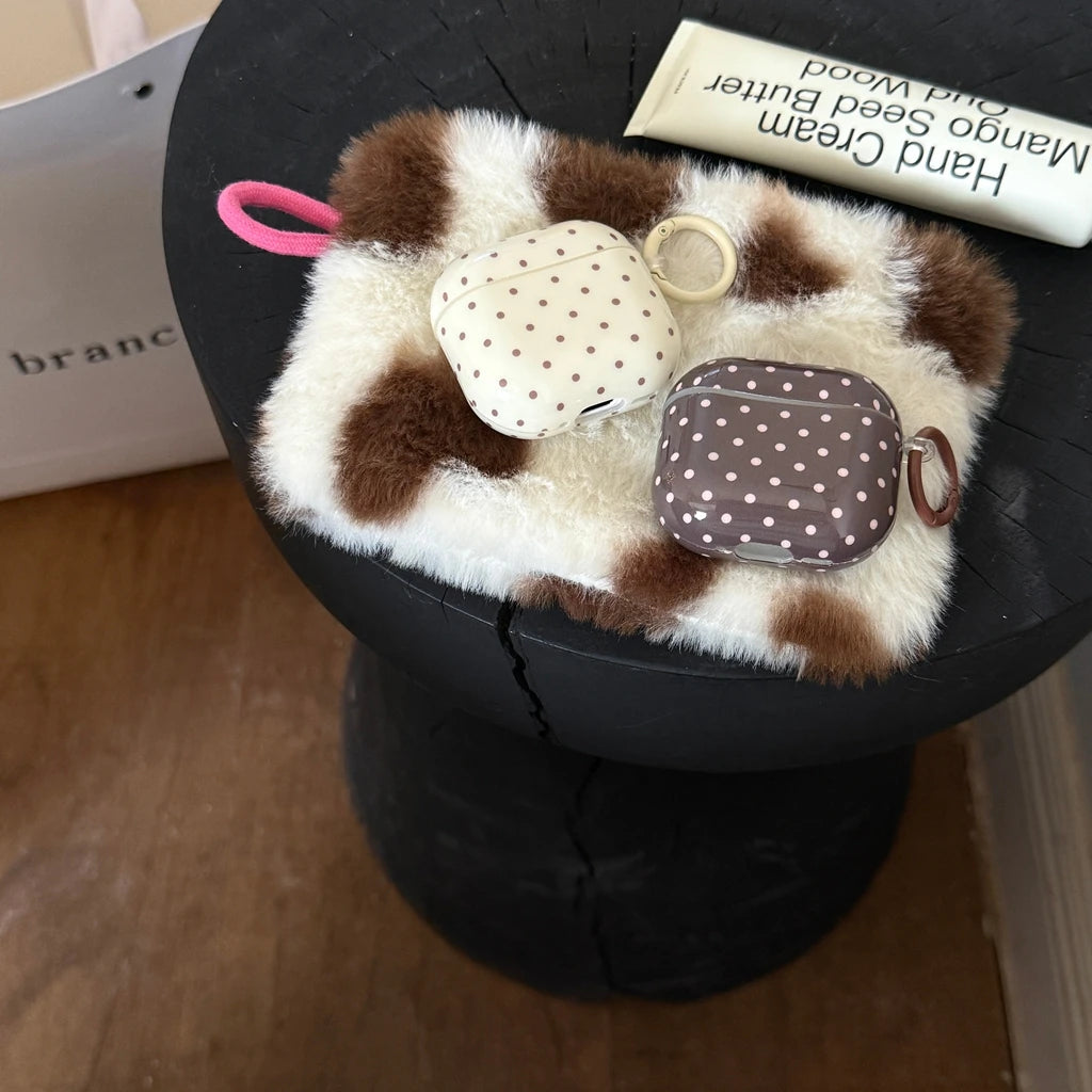 Two polka-dot AirPods Pro cases, one cream and one brown, on a fluffy cow-print pouch.