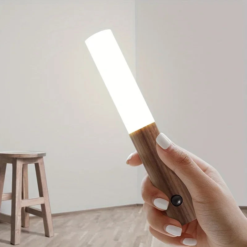 A hand holding a rechargeable LED night light with a wood-effect handle, motion sensor button, and glowing white top.