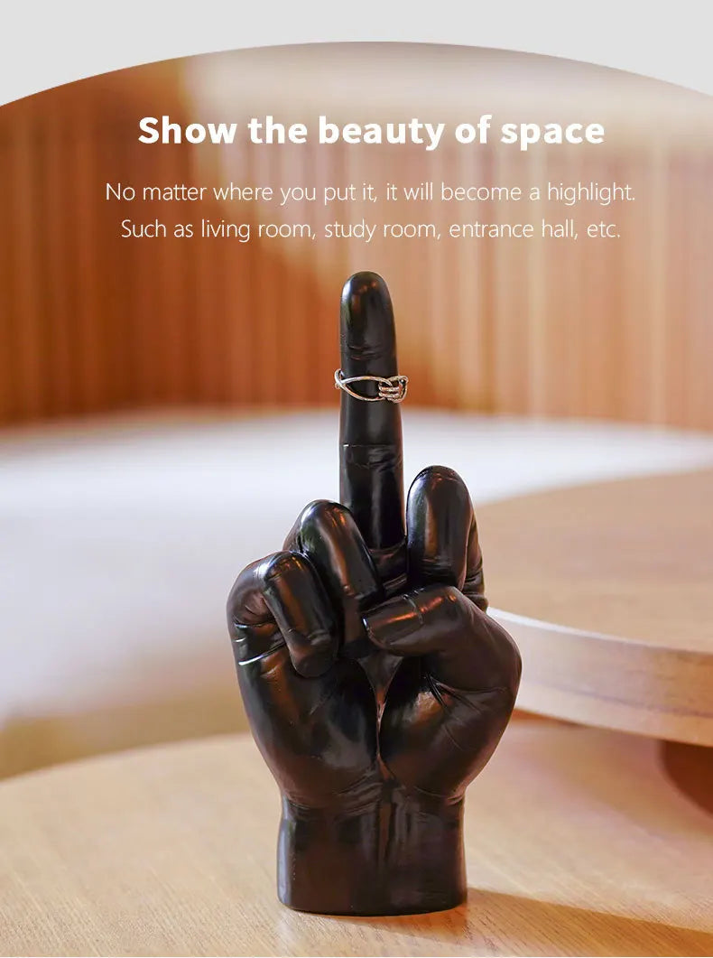 Black hand-shaped jewellery display stand with a middle finger gesture, shown holding a silver ring on a wooden table.