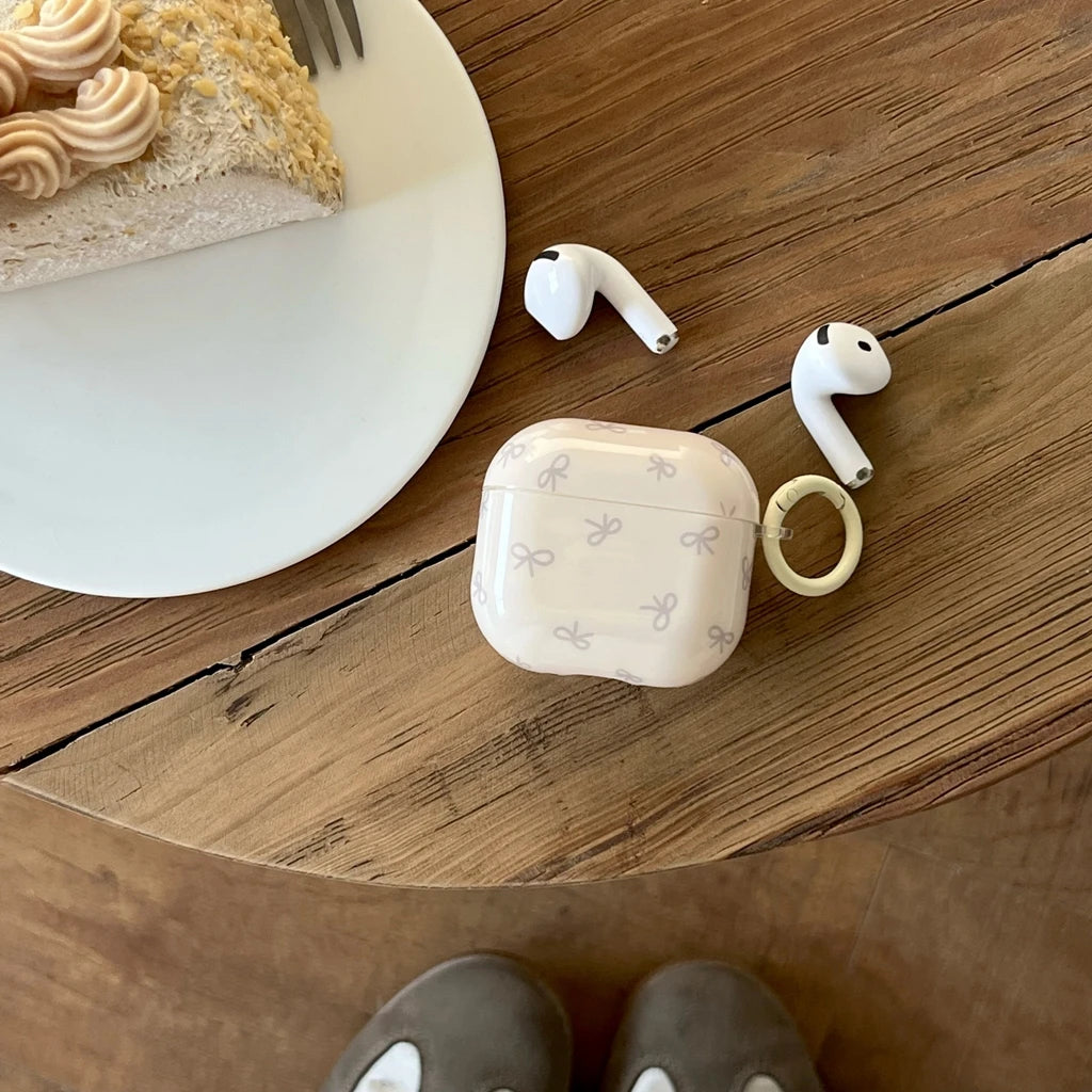 Cream AirPods case with grey bow pattern and ring attachment, shown on a wooden table.