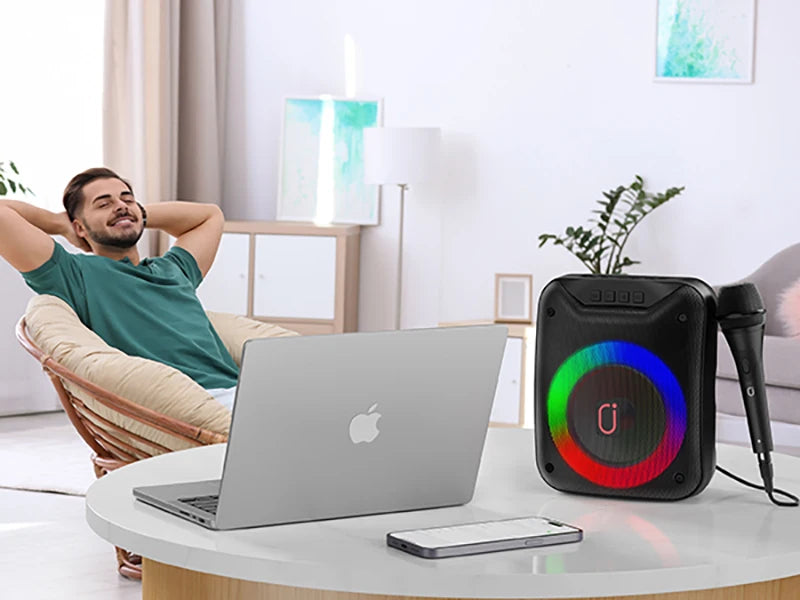 Portable black wireless speaker with multicoloured LED light ring and microphone on a table in a modern living room.