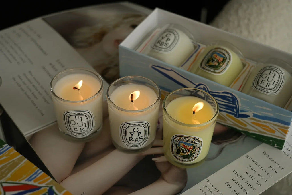 Set of three scented candles in clear glass jars, featuring glowing wicks and illustrated labels, inside a gift box.