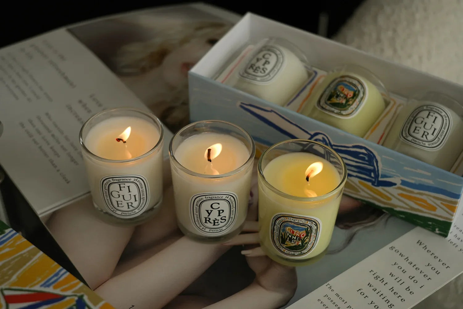 Set of three scented candles in clear glass jars, featuring glowing wicks and illustrated labels, inside a gift box.