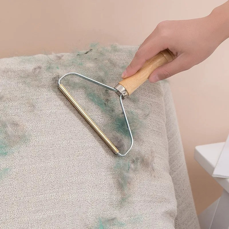 Hand using a wooden-handled fabric shaver to remove lint and pet hair from a beige textile surface.