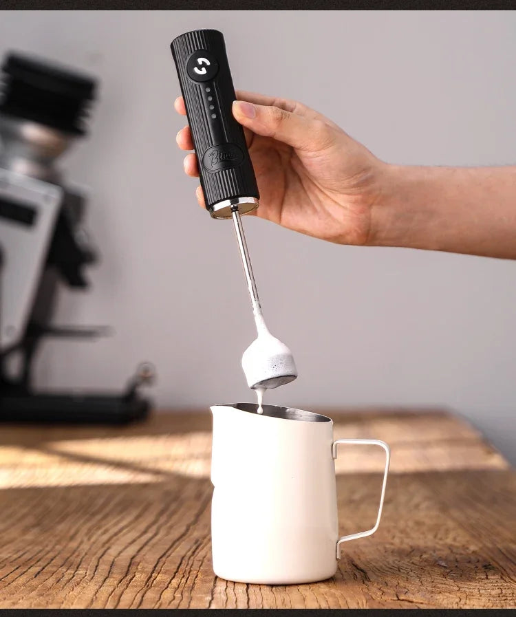 Handheld black electric milk frother with a metal whisk, held over a white stainless steel milk pitcher.