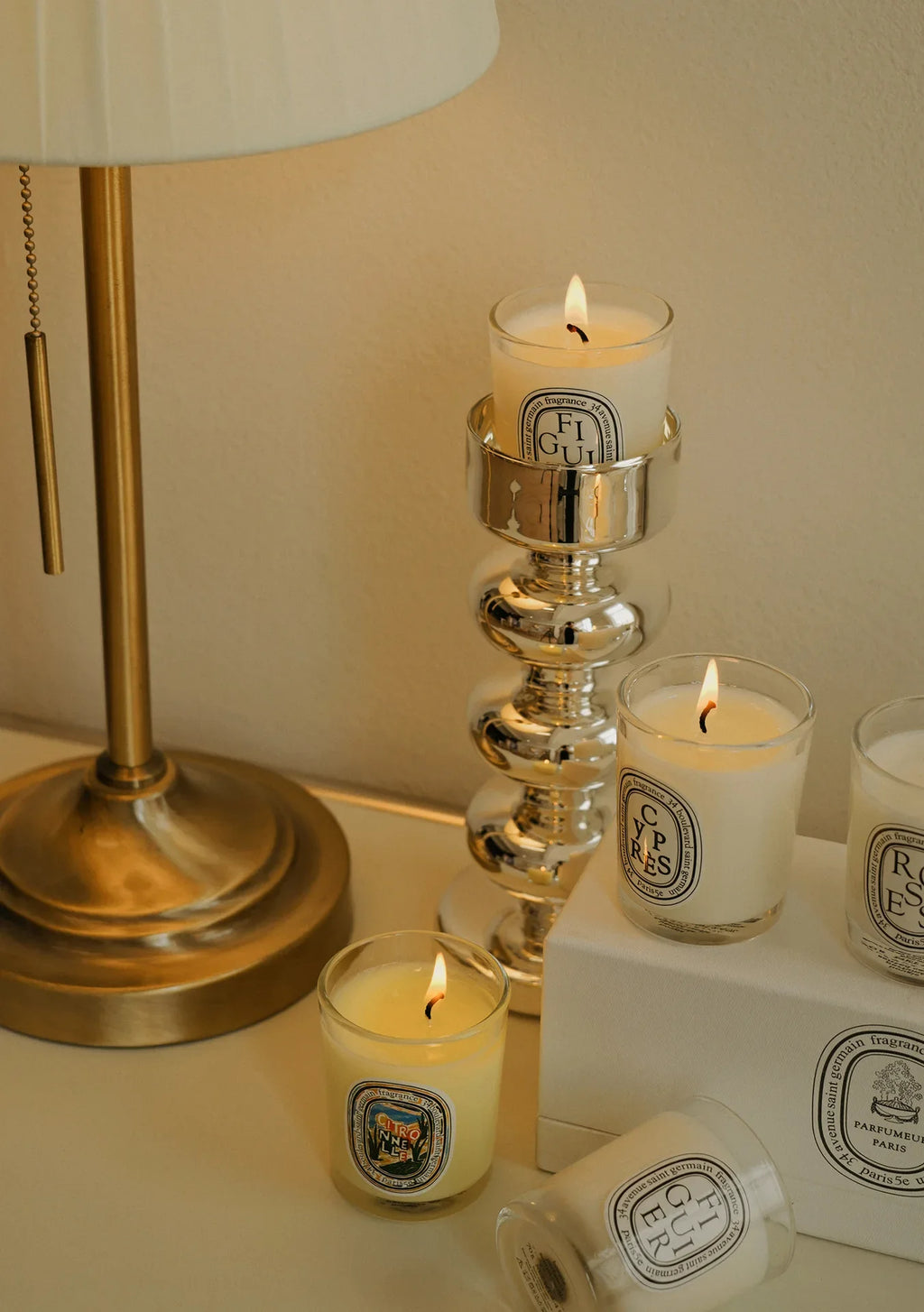 Lit scented candles in glass jars arranged on a table with a brass lamp and decorative silver candle holder.
