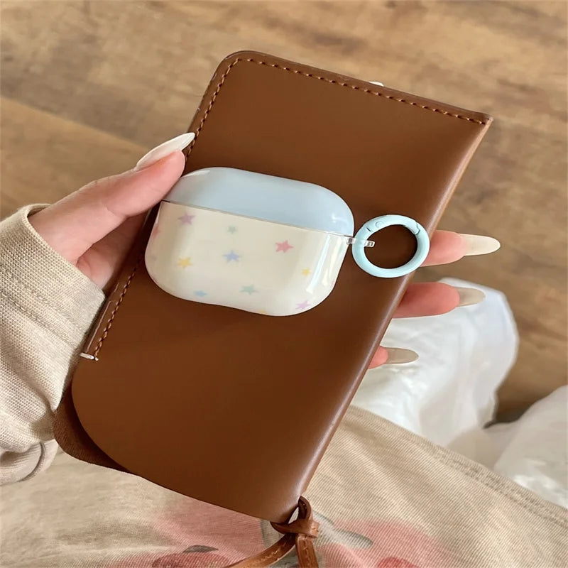 Cream and blue AirPods Pro case with pastel star pattern and matching ring clip, held against a brown leather pouch.
