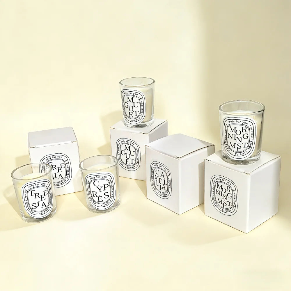 Selection of scented candles in clear glass jars with oval labels and matching white gift boxes on a cream background.