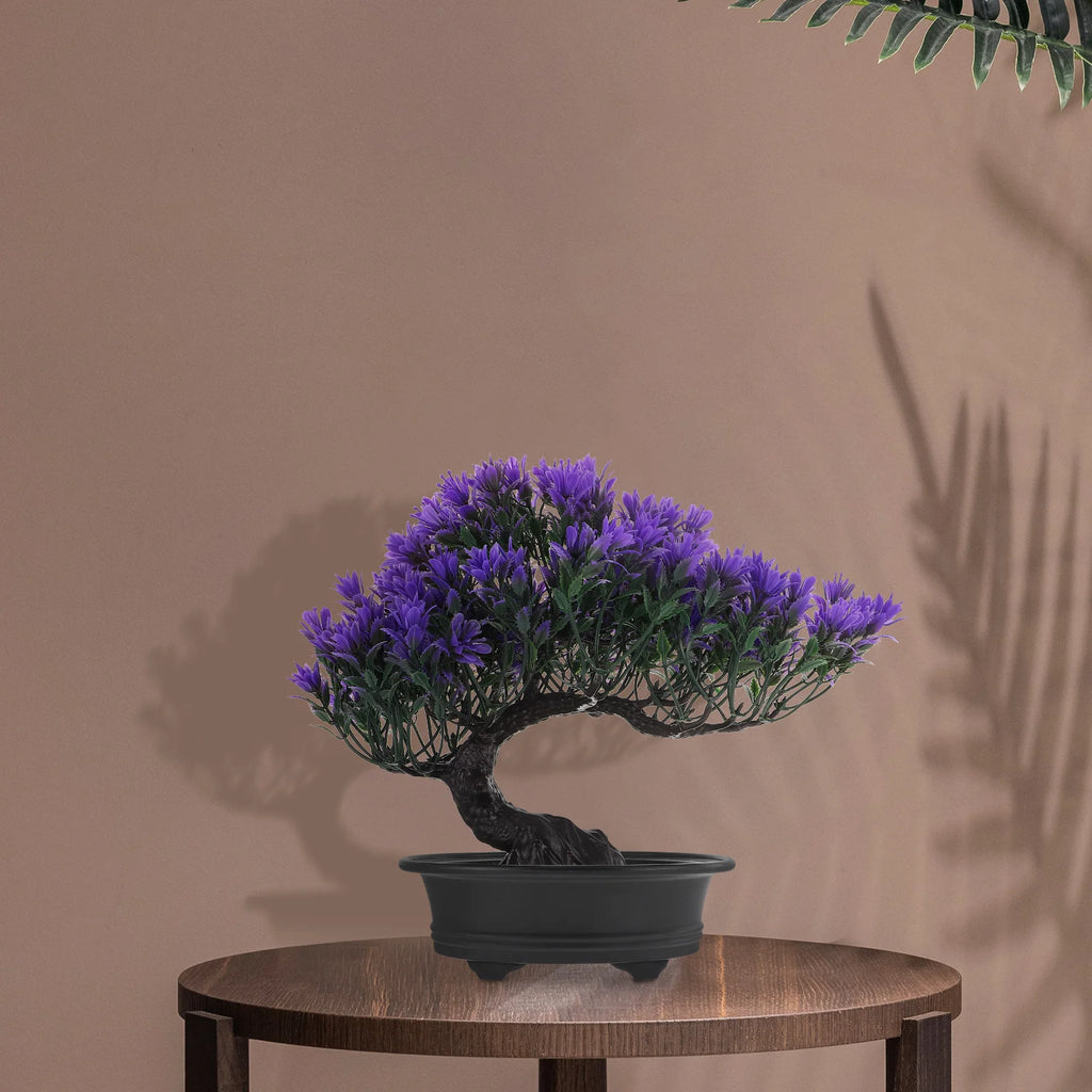 Artificial bonsai tree with vibrant purple flowers in a black pot, displayed on a wooden table against a brown wall.