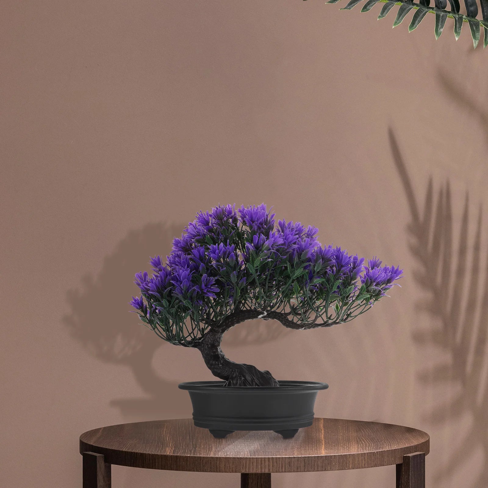 Artificial bonsai tree with vibrant purple flowers in a black pot, displayed on a wooden table against a brown wall.