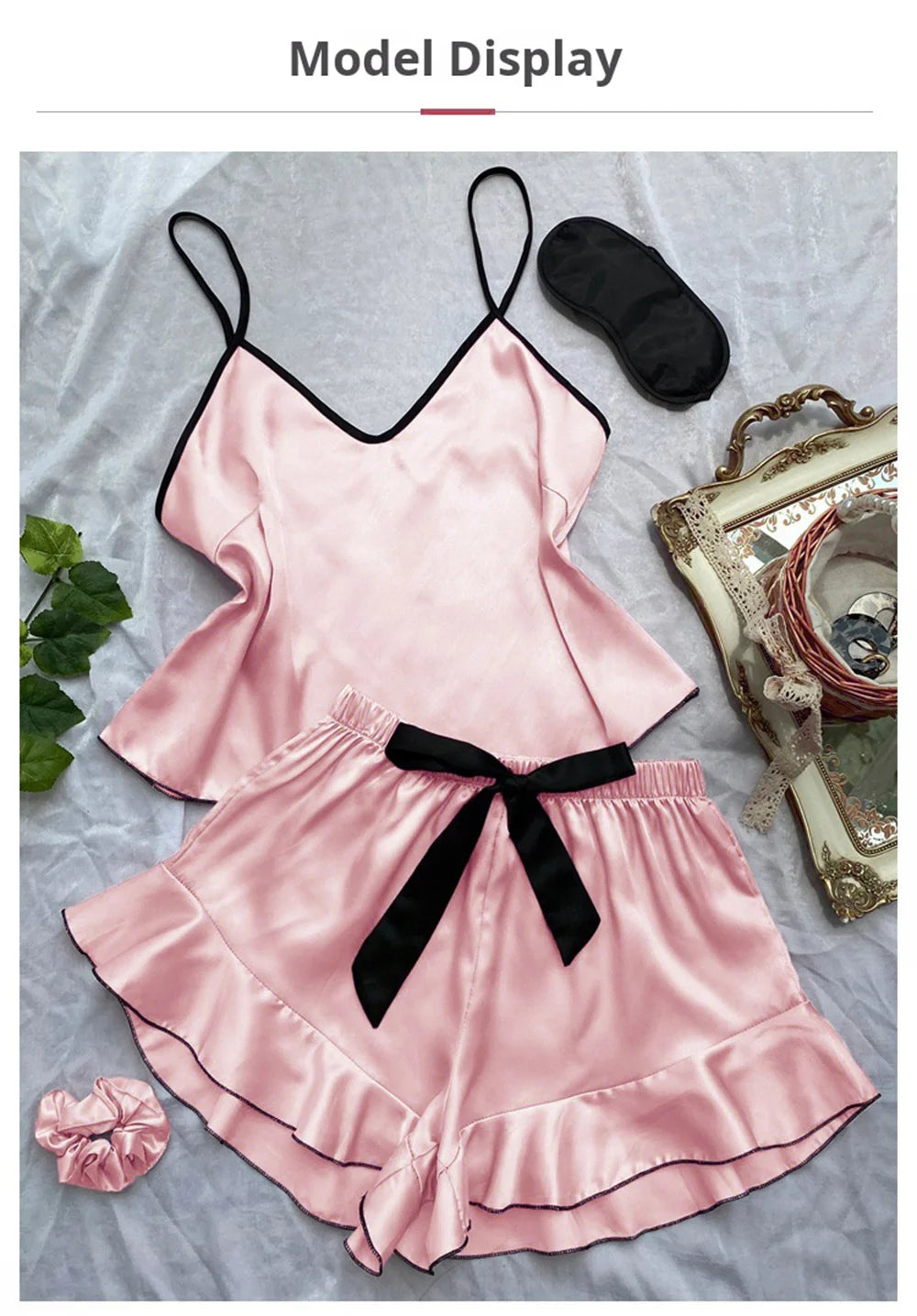 Pink satin pyjama set with camisole and ruffled shorts, featuring black trim, a waist bow, and matching sleep mask.