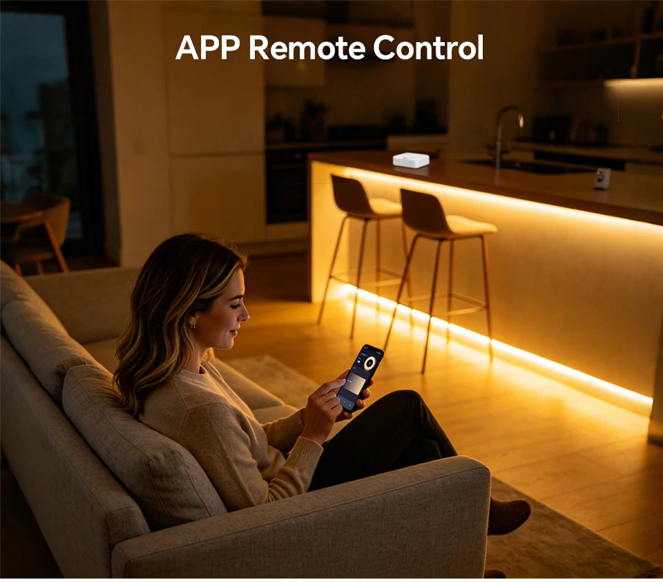 A woman sits on a sofa, using a smartphone app to control warm-toned LED strip lighting installed under a kitchen island.