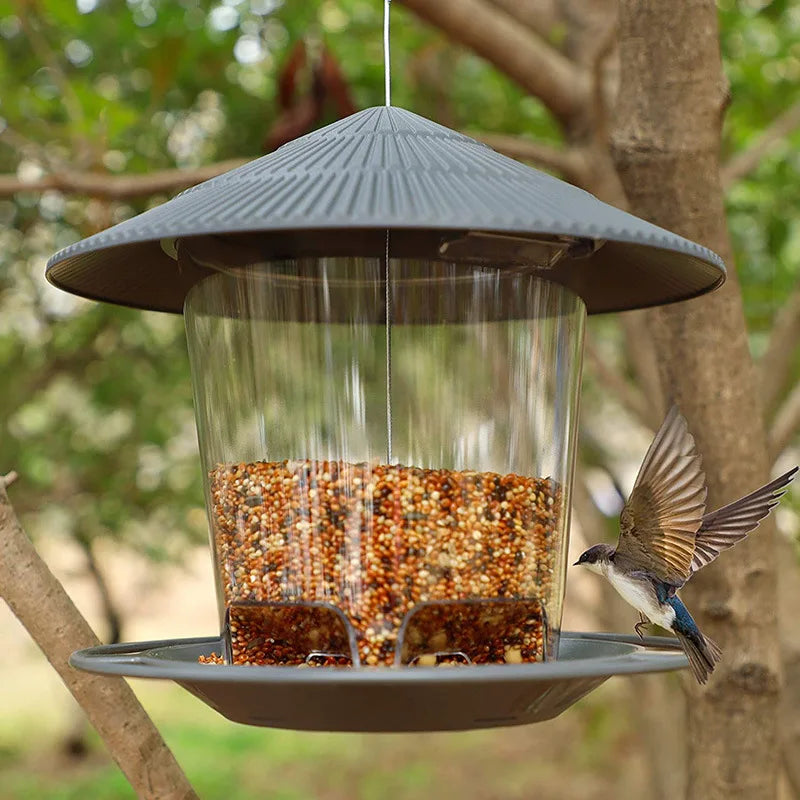 Hanging bird feeder with clear container, grey roof and base, filled with birdseed, with a bird flying nearby.