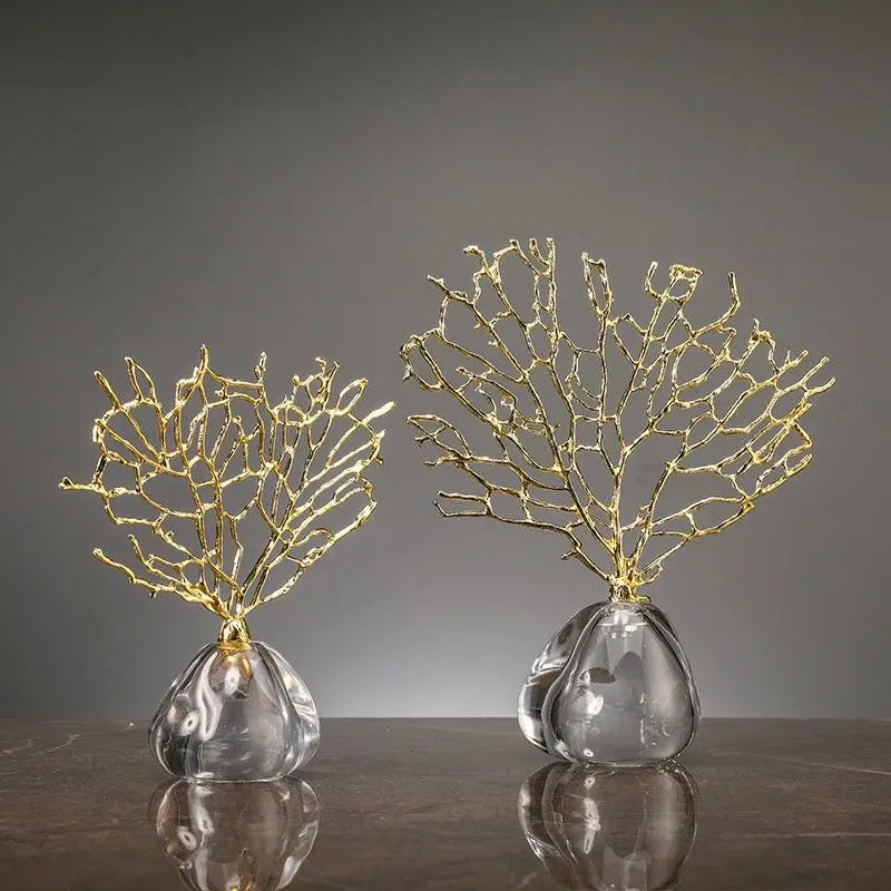 Two decorative coral-like gold sculptures on clear glass bases stand on a dark surface against a grey background.