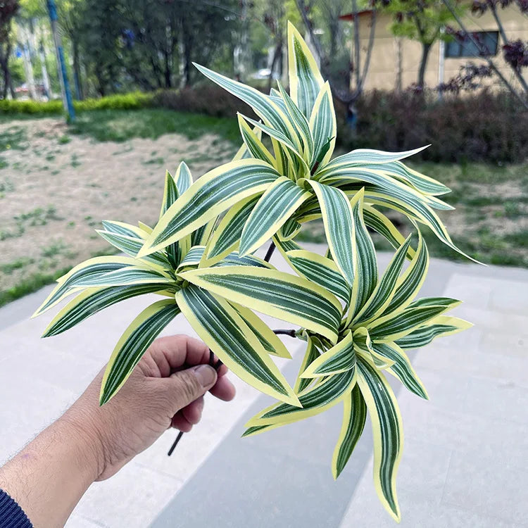 A hand holds an artificial plant branch with pointed, striped green and yellow leaves against an outdoor background.