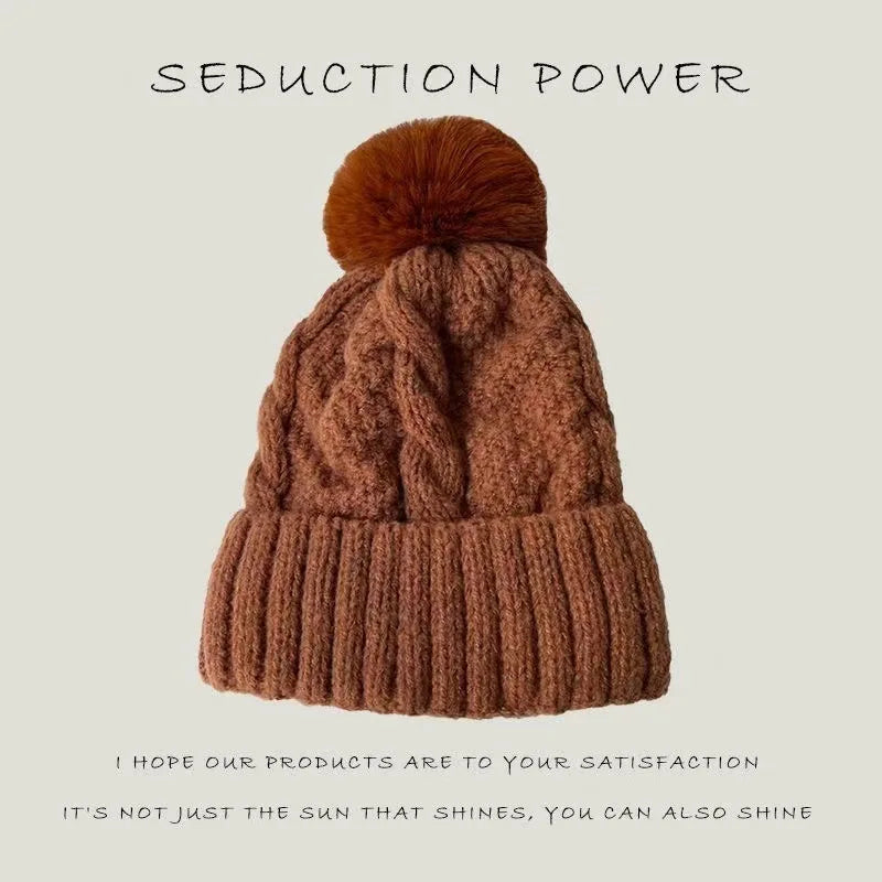 Rust brown knitted beanie hat featuring a cable-knit pattern, a ribbed turn-up hem and a matching faux-fur pom pom.