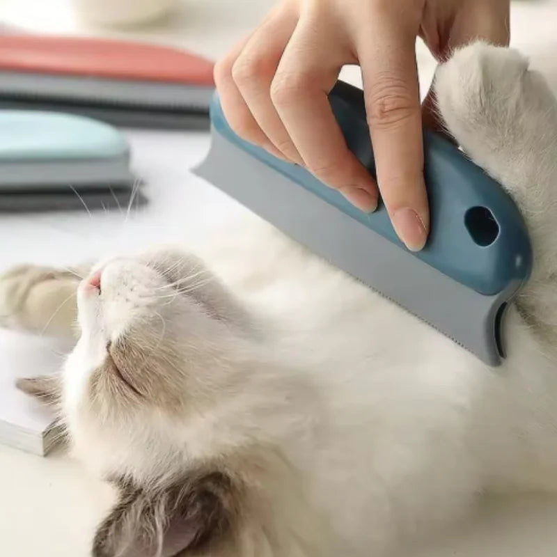 A person uses a handheld dark blue and grey pet grooming tool to brush a fluffy white cat lying on its back.