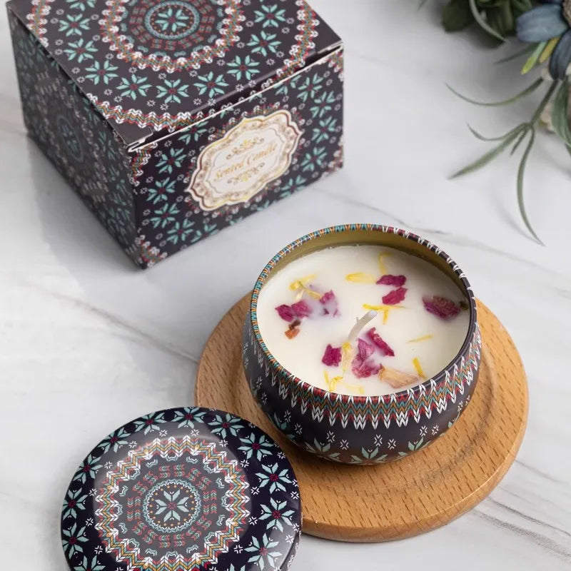 Scented candle in a patterned metal tin with floral wax, matching lid, and decorative gift box on a wooden coaster.