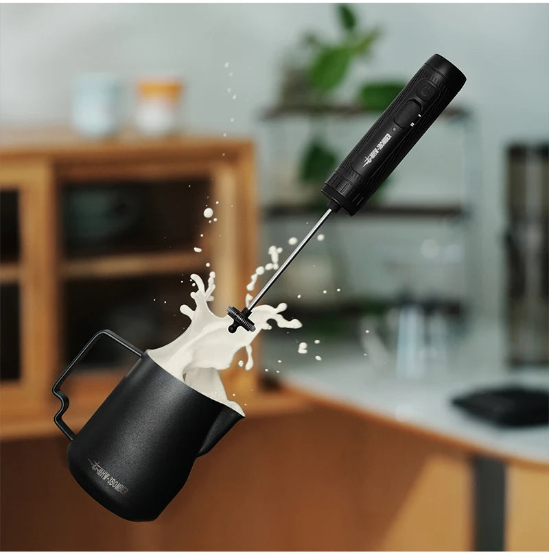 Handheld electric milk frother and matte black metal pitcher with milk splashing out. Modern, sleek kitchen tools.