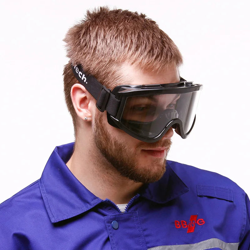 Black safety goggles with clear lenses and an adjustable strap, worn by a man in a blue work uniform.