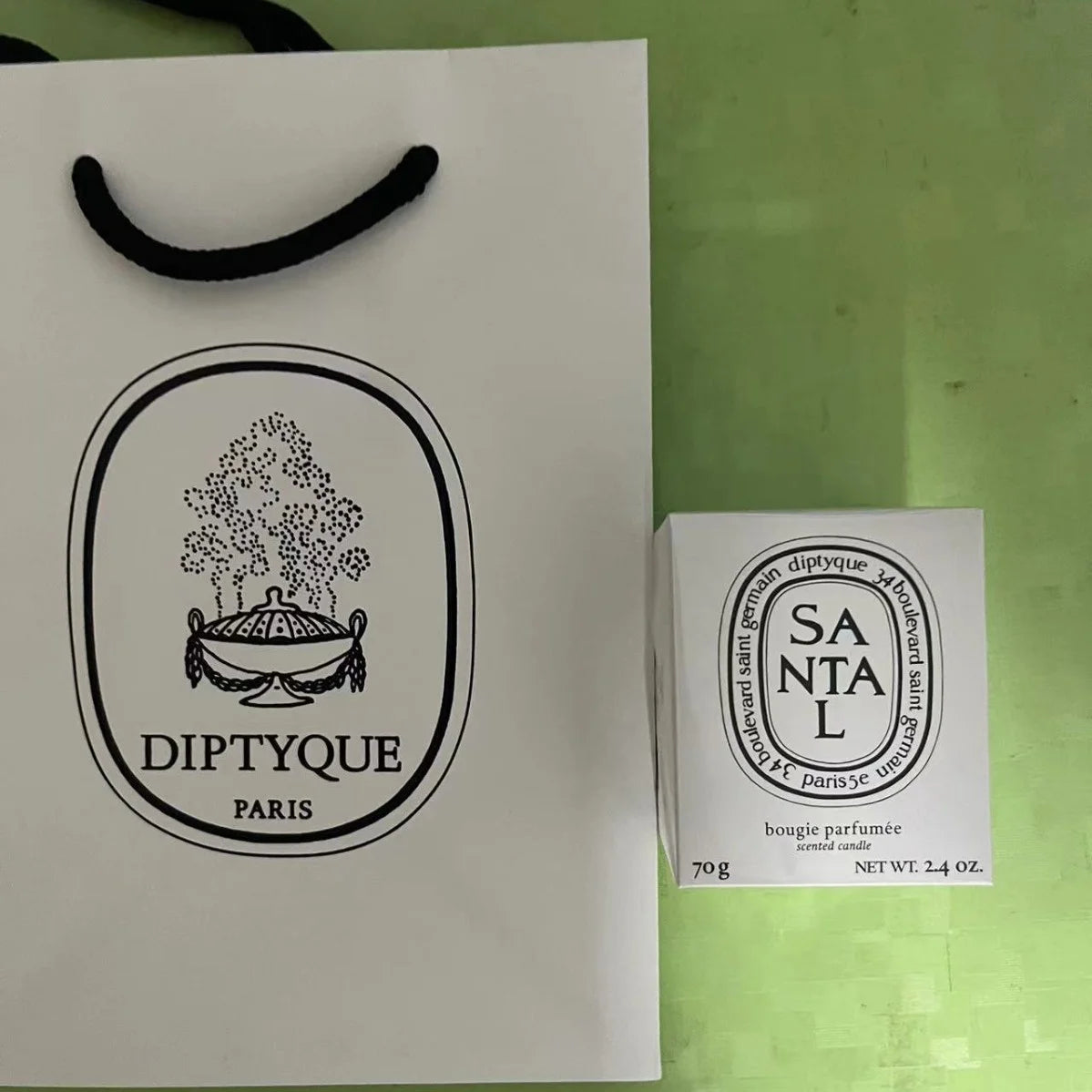 White 70g scented candle in a box next to a matching white paper gift bag with black branding.