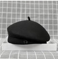 Classic black wool felt beret with a small stem top and a metallic letter 'M' logo detail on the front.