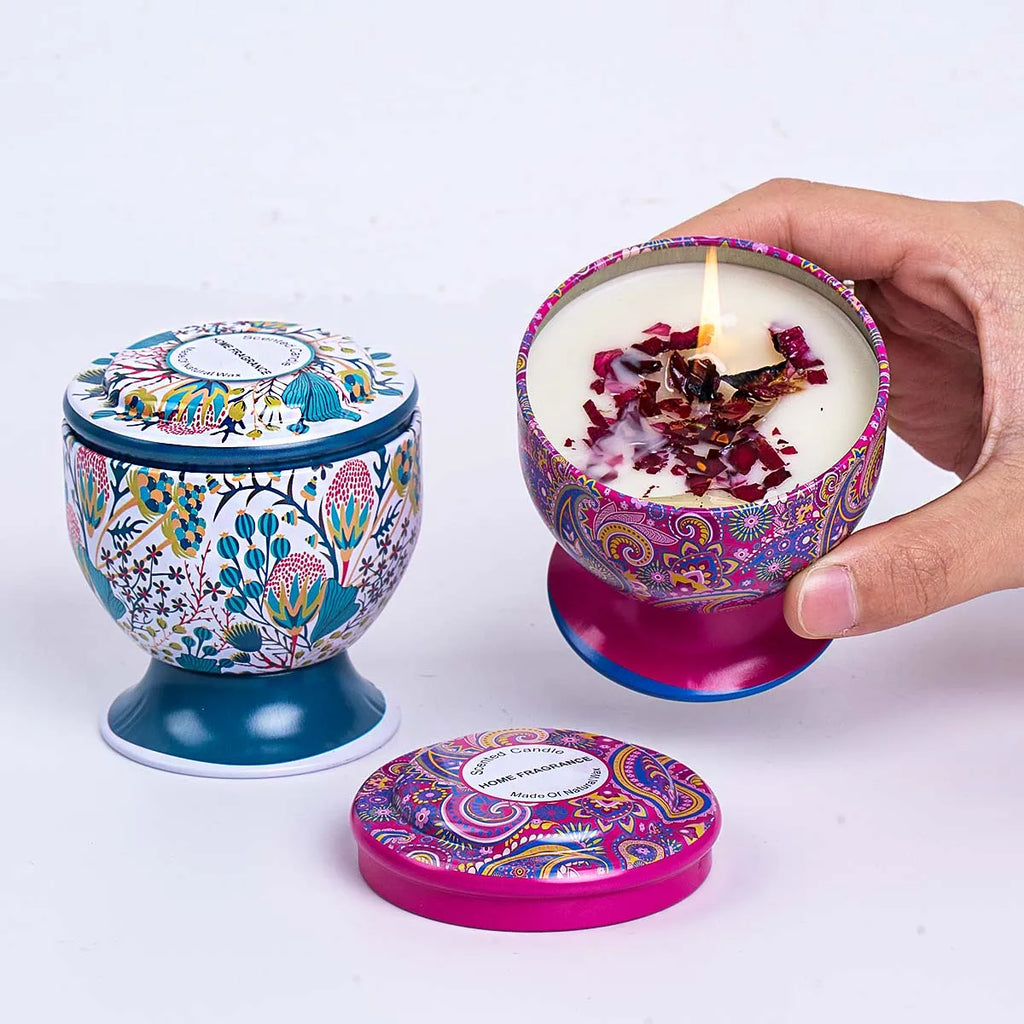 Lit scented candle in a decorative, patterned pedestal tin with dried flower petals on wax and matching lids.