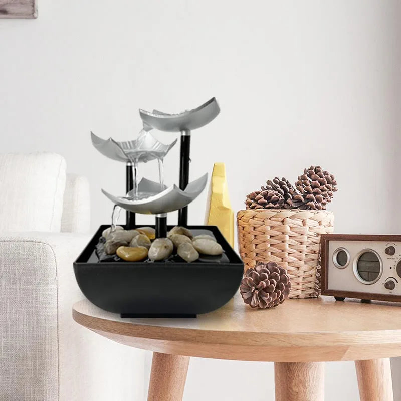 Modern tabletop water fountain with three silver tiered bowls, black pillars, and river stones in a black base.