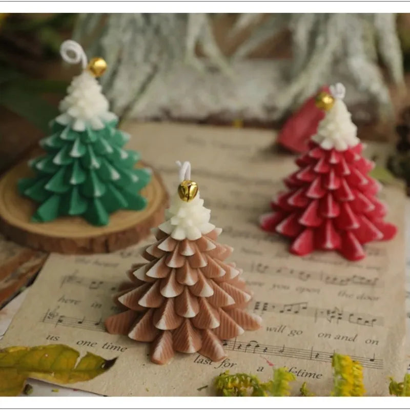 Three Christmas tree-shaped wax candles in green, red, and brown, featuring white tips and a small gold bell detail.