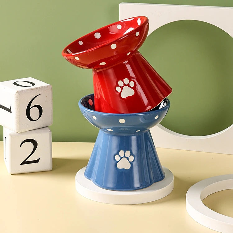 Two stacked, elevated ceramic cat bowls in red and blue, featuring a white polka dot pattern and a paw print motif.
