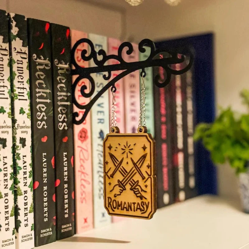 Black scrollwork wall bracket holding a wooden sign engraved with 'Romantasy' and crossed daggers, hanging on a shelf.