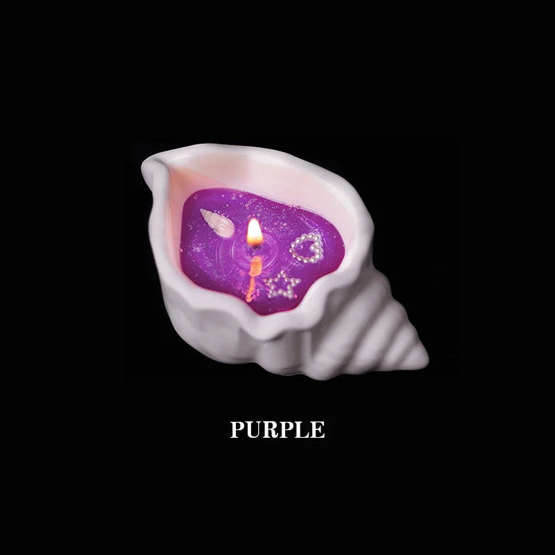 Lit purple candle in a white ceramic seashell-shaped holder with small shell, heart, and star decorations inside.
