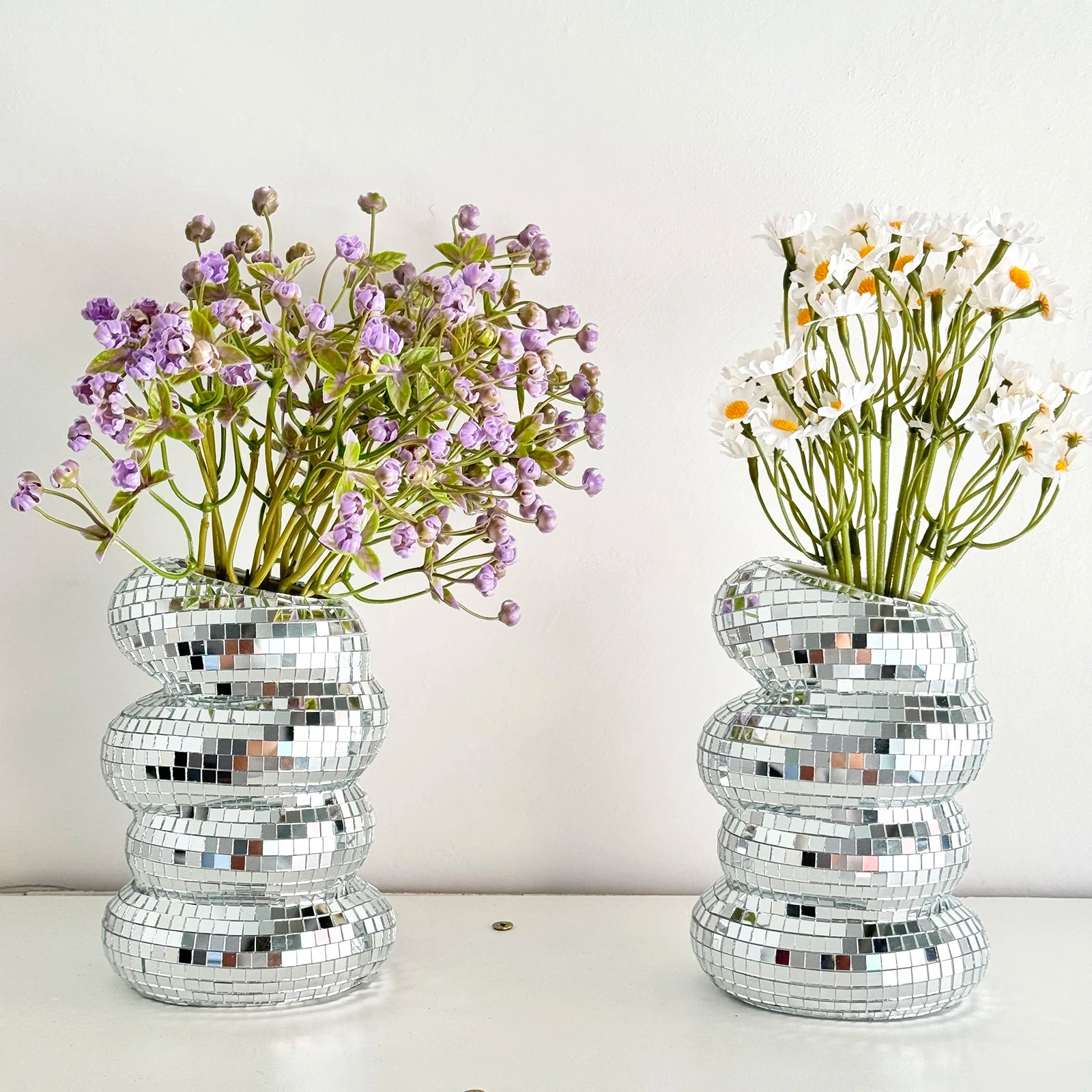 Two stacked-sphere disco ball vases on a white surface, one filled with purple flowers, the other with white daisies.