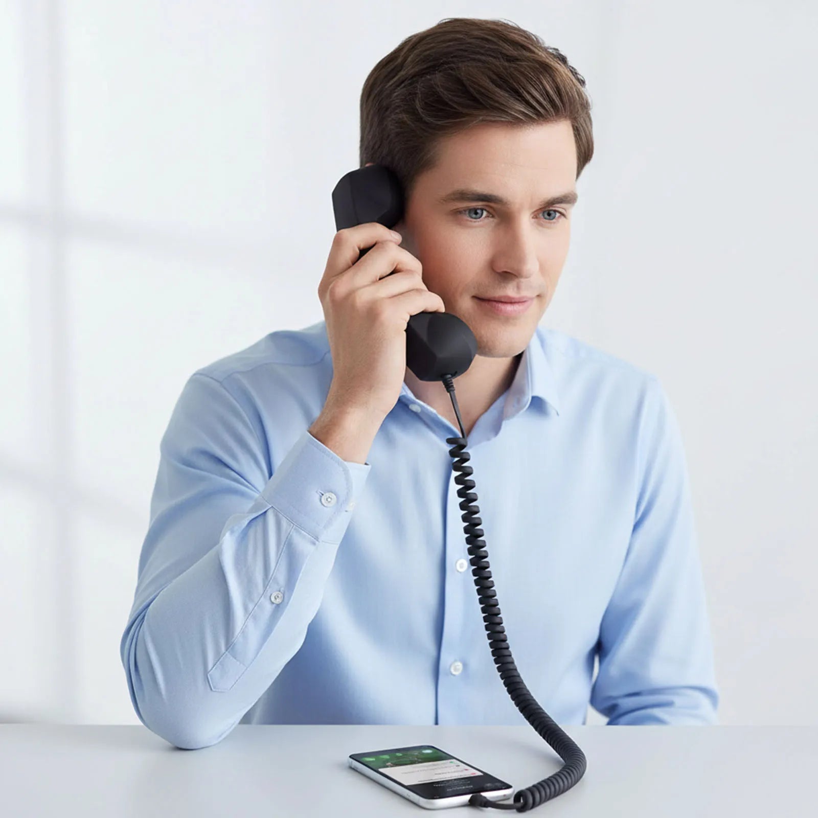 Man in a blue shirt using a retro-style black corded telephone handset connected to a smartphone.