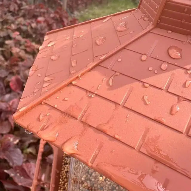 Close-up of a brown, shingled bird feeder roof with water droplets, outdoors against blurred red foliage.