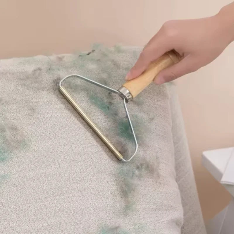A hand uses a portable metal fabric lint remover with a wooden handle to clean fluff off a beige sofa.