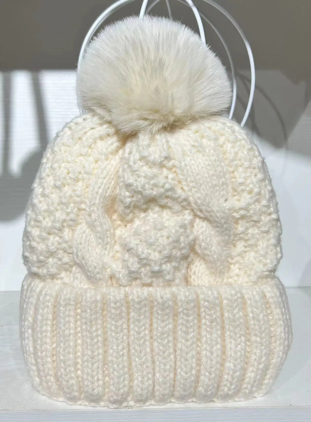 Cream cable-knit winter beanie featuring a thick ribbed turn-up hem and a matching faux-fur pom pom on top.