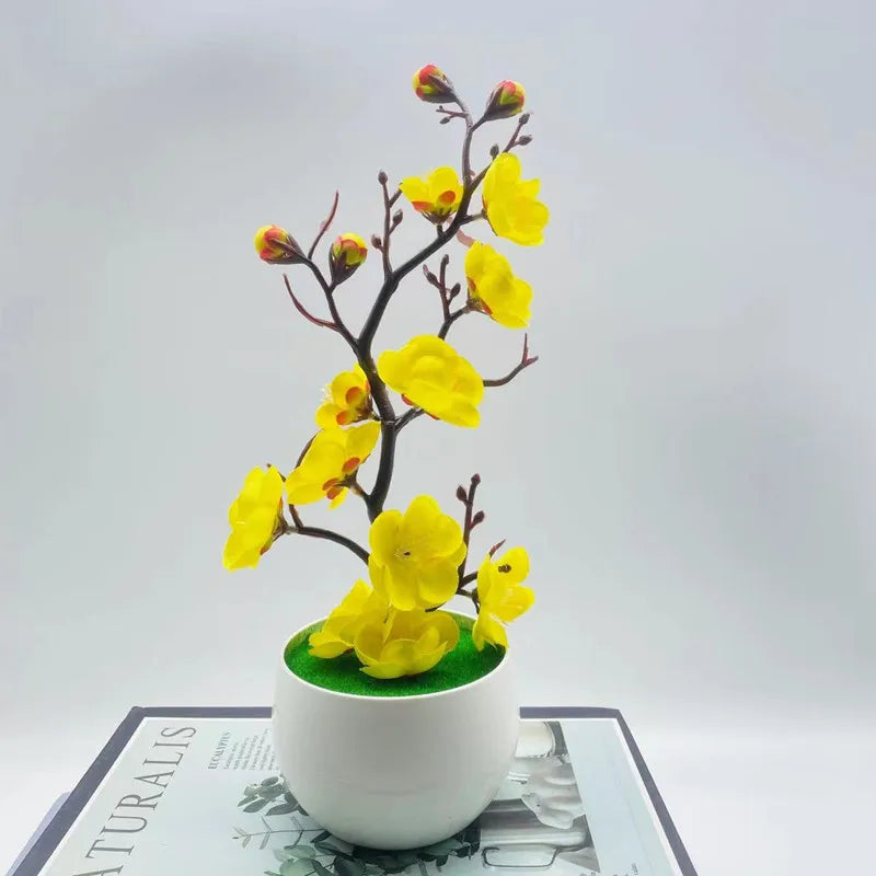 Artificial yellow plum blossom plant in a round white pot with green faux grass, set on a book.