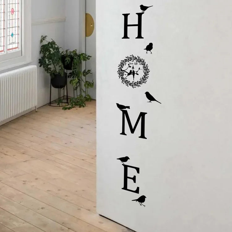 Black 'HOME' wall sticker featuring a circular floral wreath and silhouette birds, applied to a white wall.