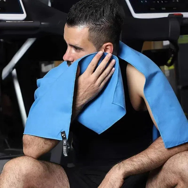 A man in a gym wearing a blue microfibre towel around his neck and wiping his face.