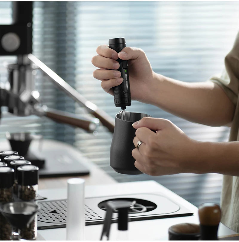 Handheld black electric milk frother being used with a matching black metal pitcher in a modern coffee station.